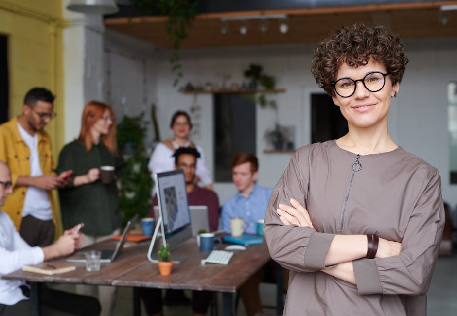 Smiling businesswoman with curly hair stands confidently in a modern office space with colleagues.