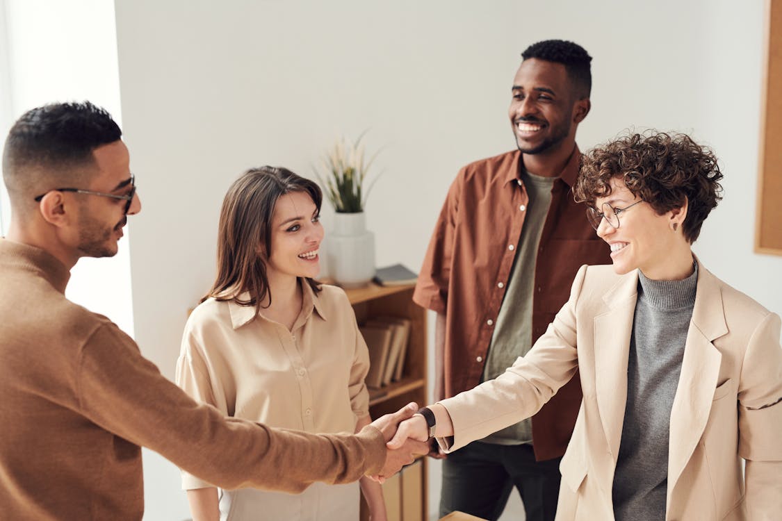 White woman with short hair shaking a black guy's hand as if they are sealing a business deal. White woman with mid length hair and black guy with watch and smile on the background. Both guys have buzzcut hair