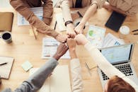 俯视图显示，一支多元化团队的拳头在会议桌上相碰，桌上摆着文书工作和笔记本电脑，象征着团队合作