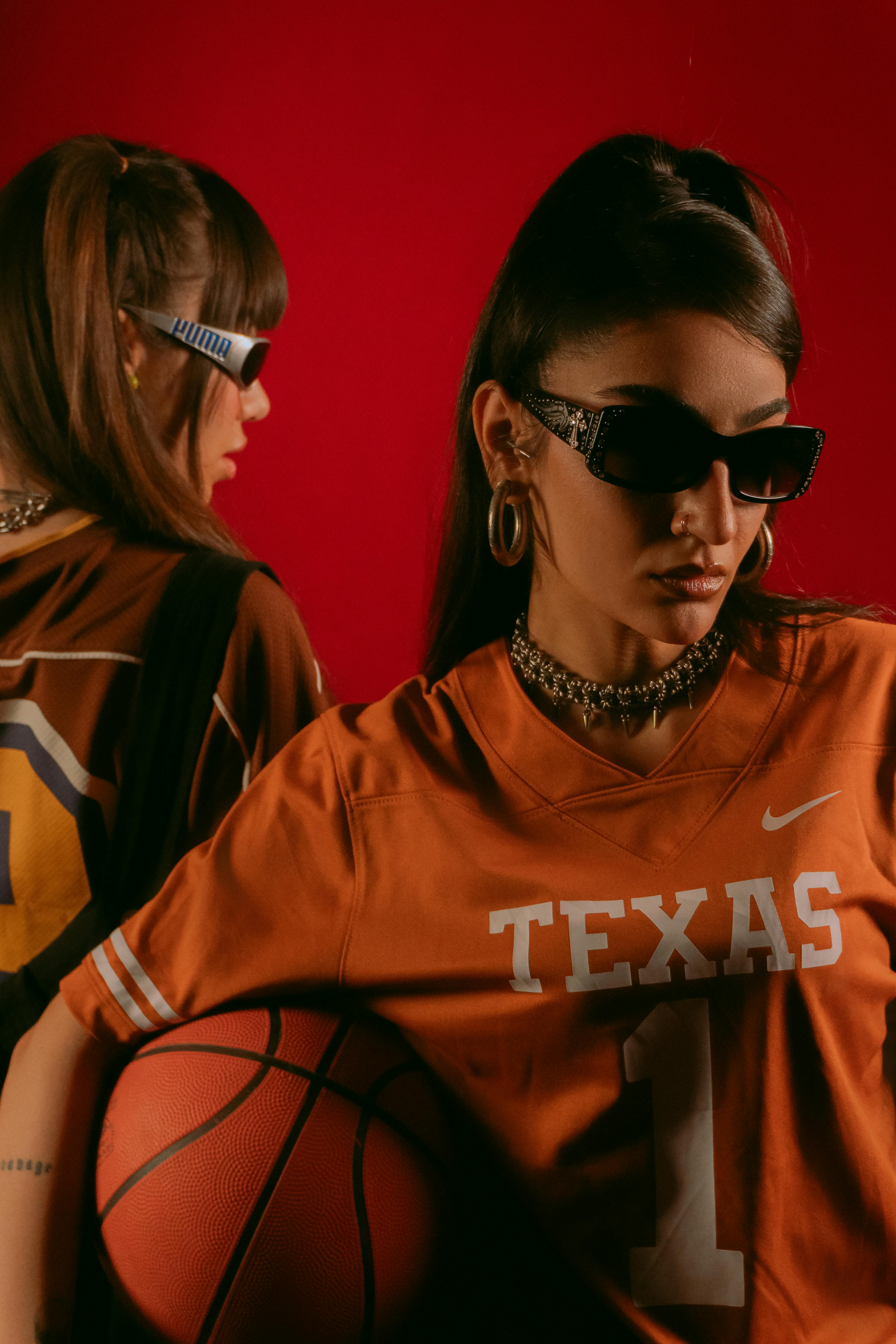 Two fashionable women in sports jerseys with sunglasses and basketball against a red backdrop.