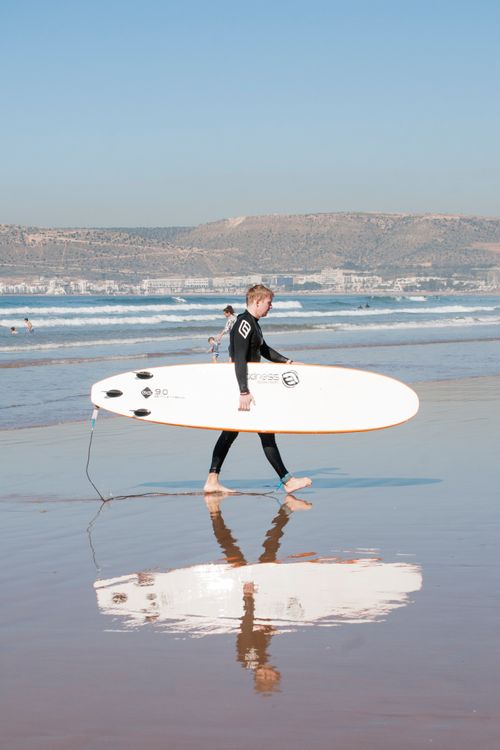 Free A surfer walks along Agadir beach carrying a surfboard, reflecting in wet sand. Stock Photo