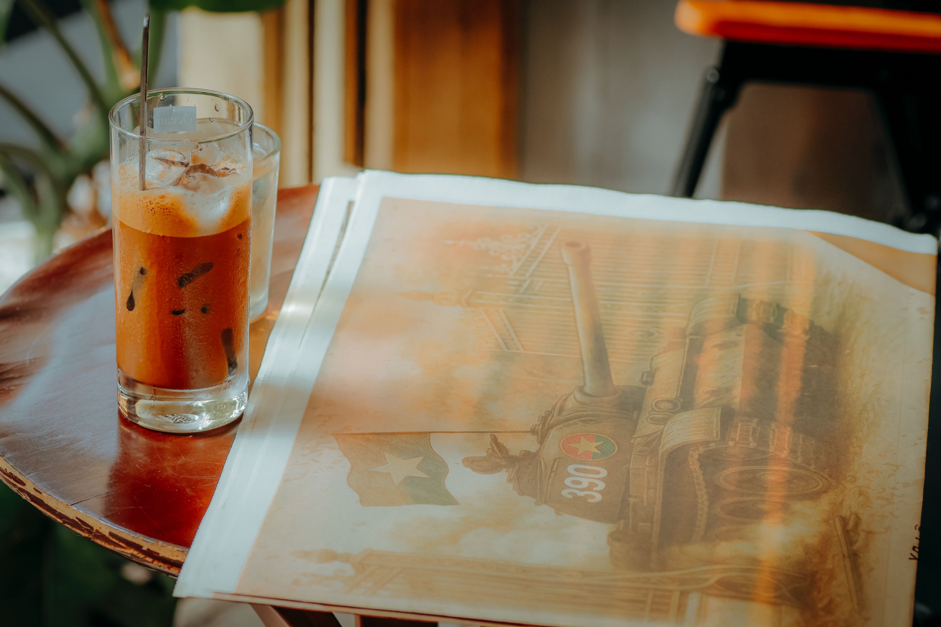 A glass of iced coffee sits on a table next to a vintage tank illustration in soft, warm light.