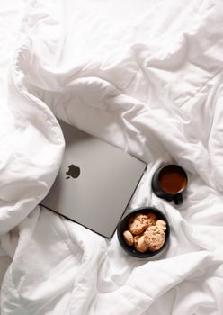 A relaxing morning scene with a laptop, coffee, and cookies on a white bed, perfect for work or leisure.