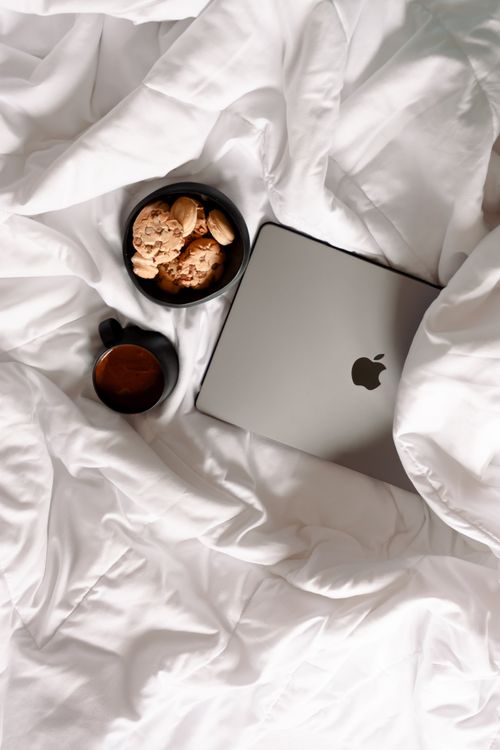 Free A laptop, cookies, and coffee on a cozy white bed setting ideal for relaxed home work days. Stock Photo