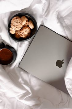 A relaxed setup featuring a MacBook, cookies, and coffee on a cozy bed with white sheets.