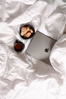 A cozy morning setup featuring a laptop, coffee, and cookies on a white bedspread. Perfect for relaxation.