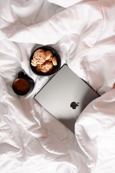 A relaxing morning setup with coffee, cookies, and laptop on soft white bedding, perfect for leisure.