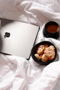 Flat lay of a laptop, coffee, and cookies on soft white bedding for a cozy work-from-home vibe.