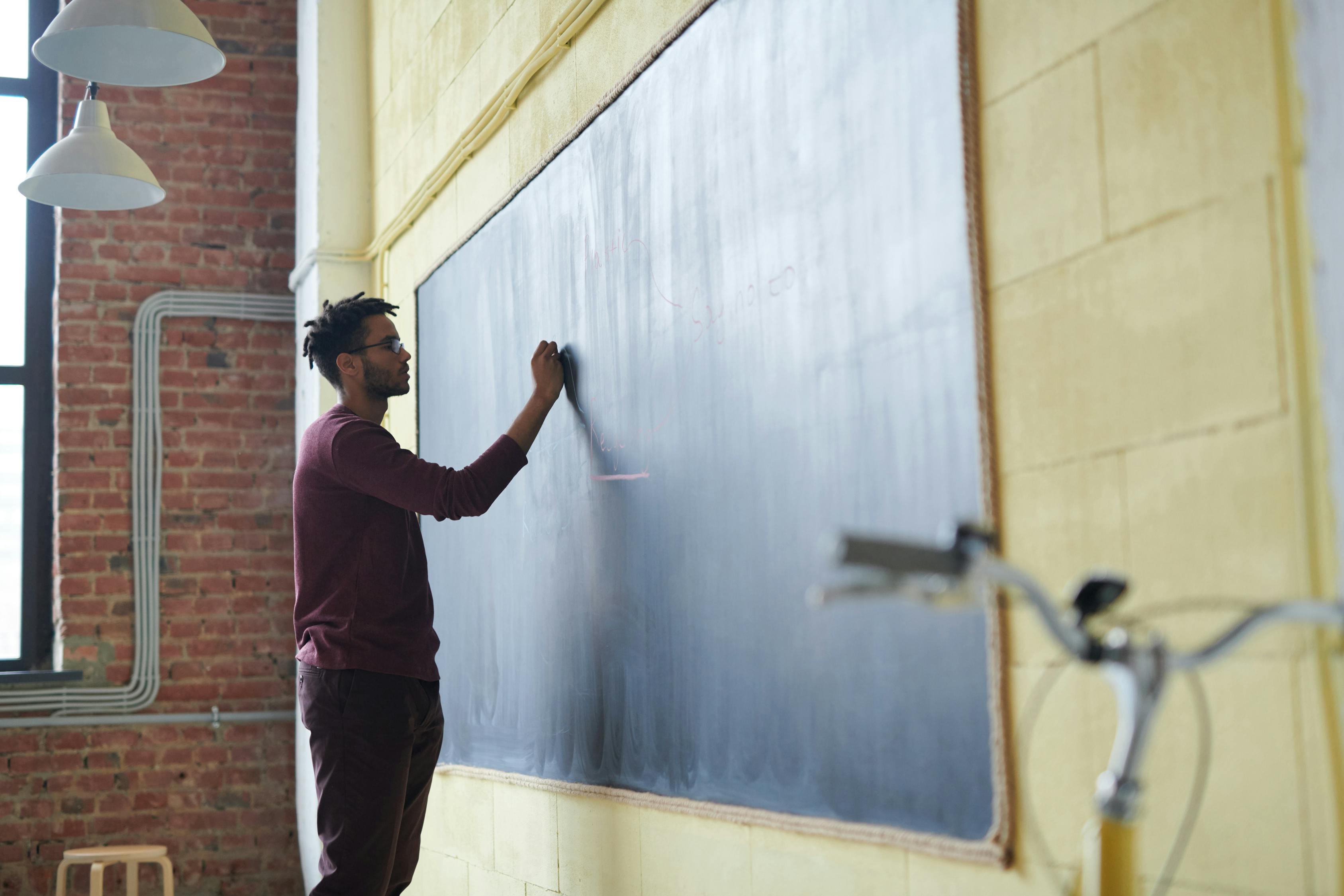 Image of a man standing before a blackboard. He is writing on the board with chalk.