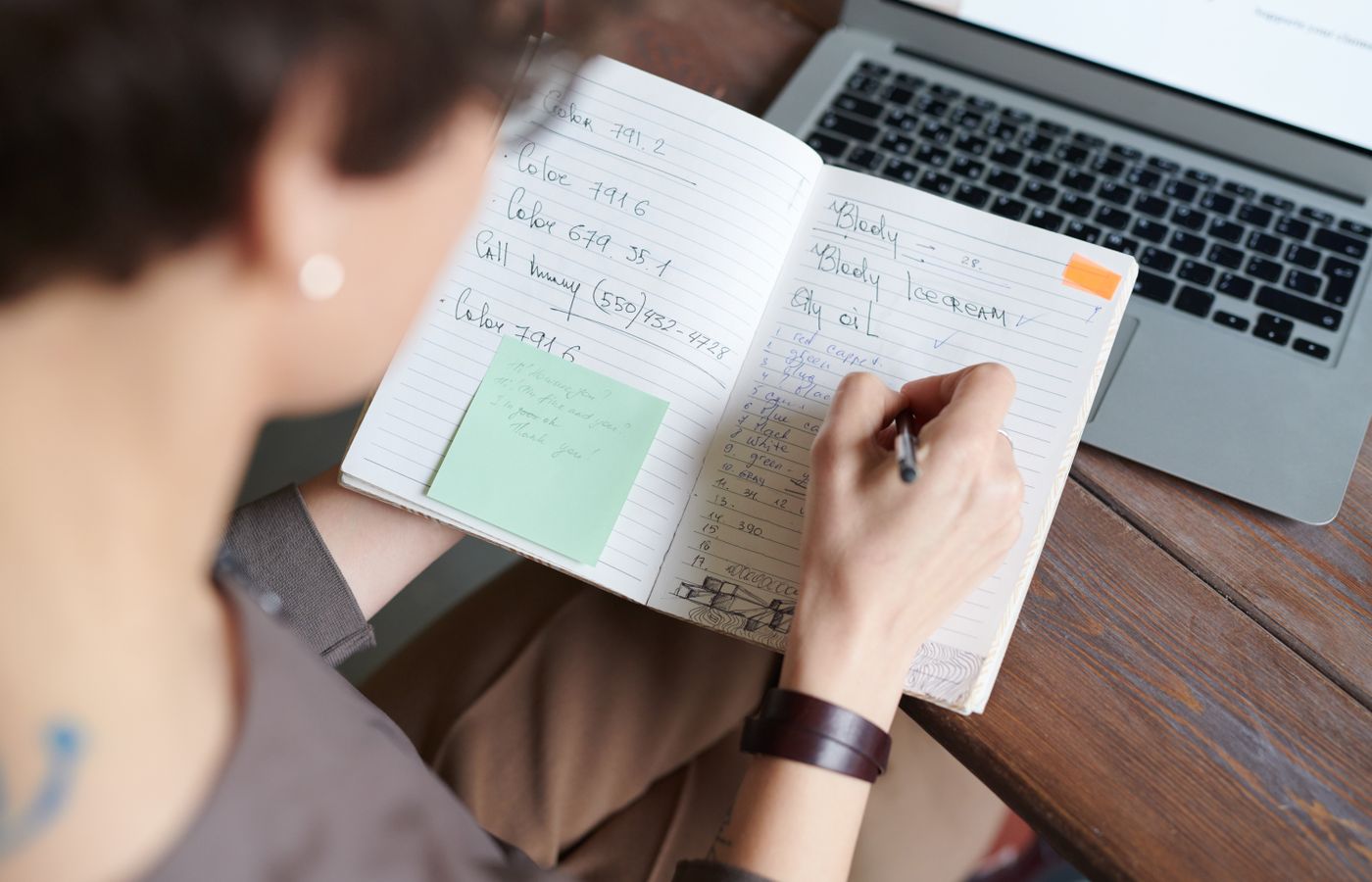 An adult writing in a notebook with a laptop on a wooden table for work or leisure.