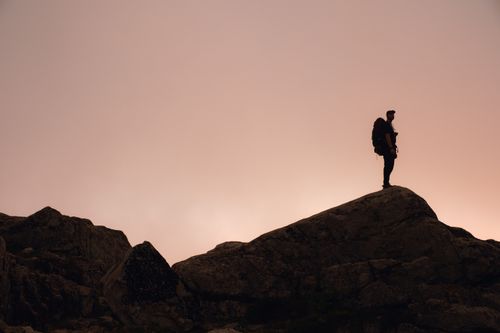 Free Silhouette of a lone hiker standing on a mountain peak against a stunning sunset sky. Stock Photo
