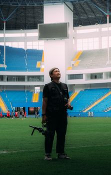 Cameraman standing on a stadium field during a sports event with vibrant seating in the background.