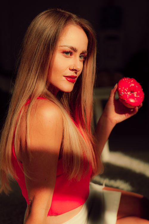 Free Beautiful woman in vibrant red top holds a pink donut, basking in warm sunlight. Stock Photo