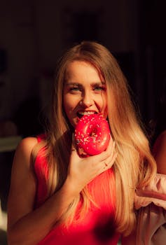 Blonde woman in a pink shirt joyfully biting into a vibrant pink donut.