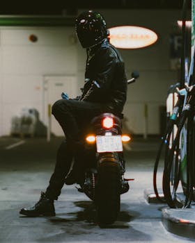 A motorcyclist in black gear refueling at a gas station during nighttime, creating a mysterious and cinematic vibe.