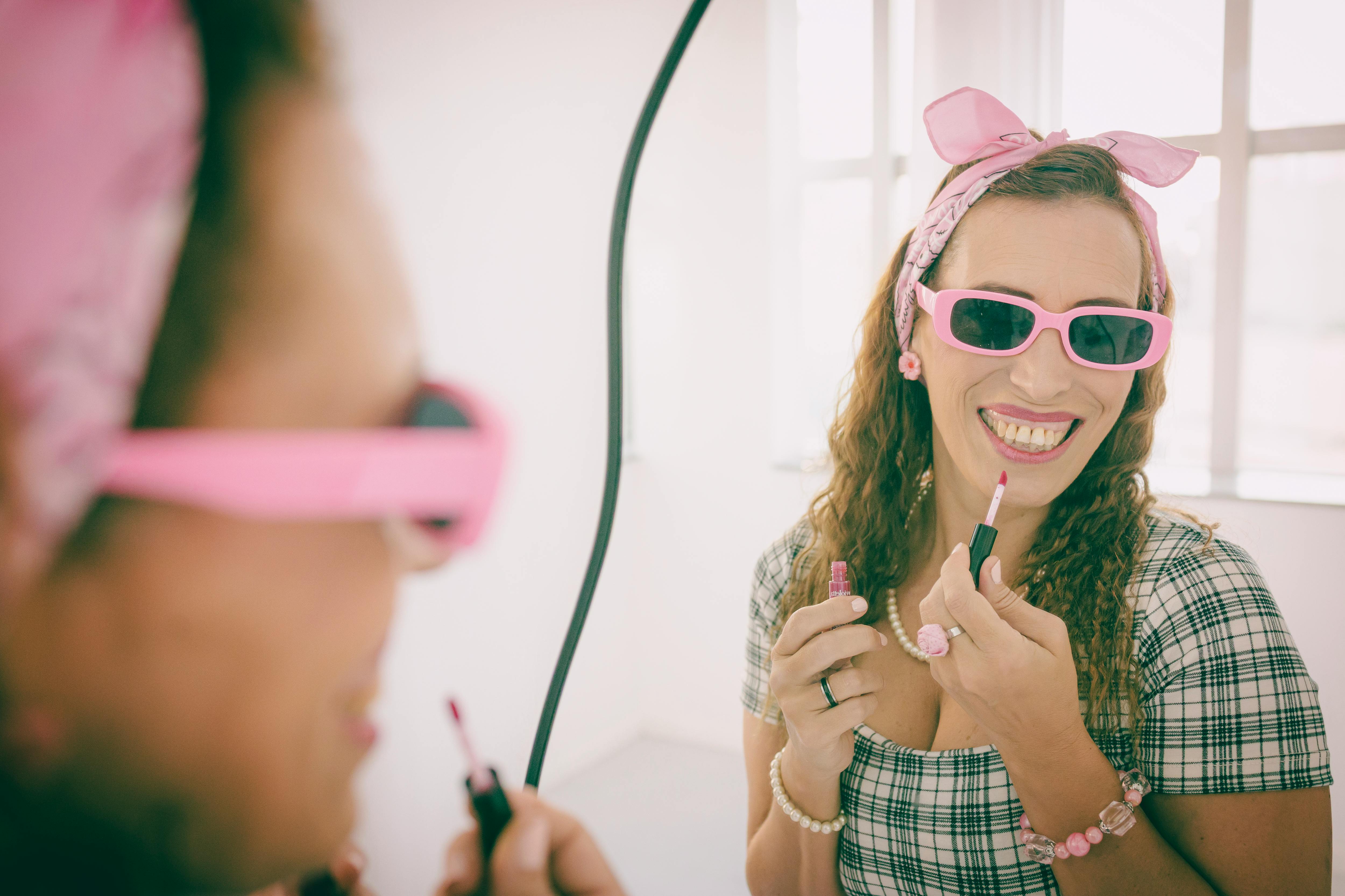 Free Cheerful woman in pink accessories applies lipstick in front of a mirror, exuding a vintage vibe. Stock Photo