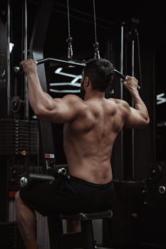 Muscular man performing a lat pulldown exercise in a dimly lit gym for strength training.