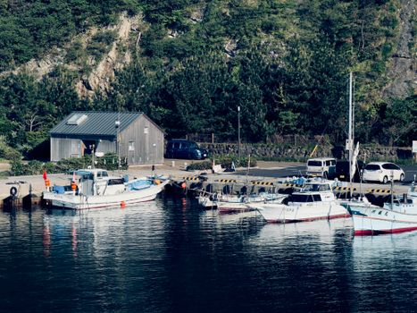 Tranquil small harbor with boats and a boathouse, framed by lush greenery and rocky cliffs.