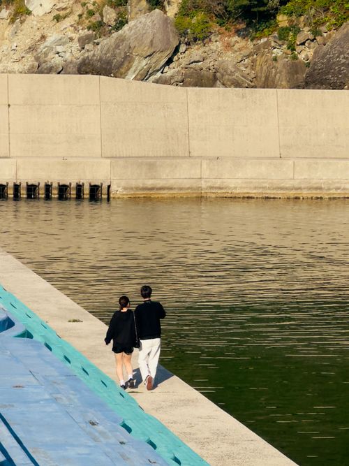 Free Two people walking by a harbor wall under clear skies. Tranquil seaside moment. Stock Photo