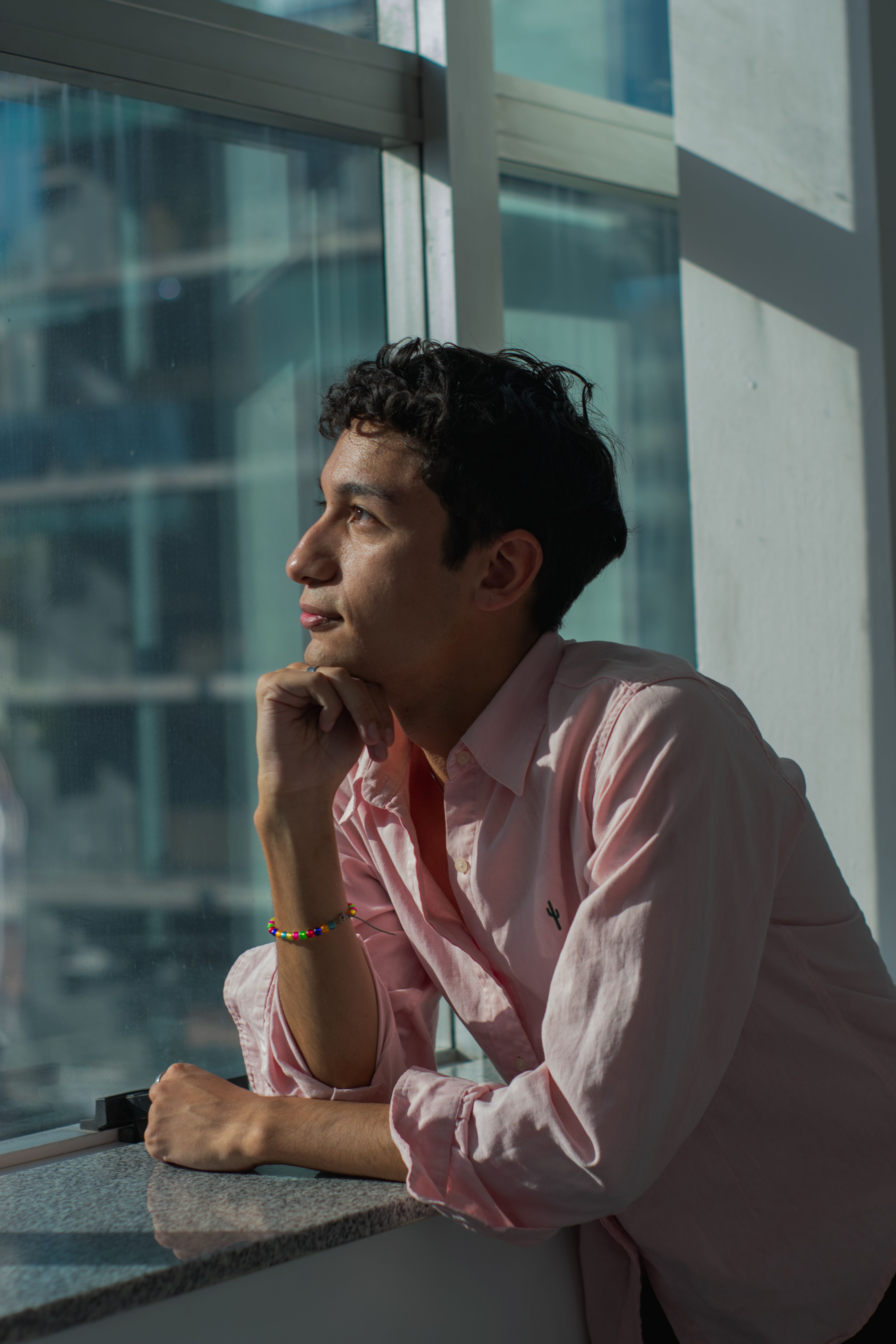 Young adult in pink shirt thoughtfully gazing out the window, capturing a moment of introspection indoors.