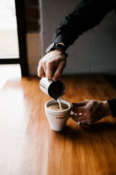 Barista elegantly pouring milk into a coffee cup on a wooden table, indoors.