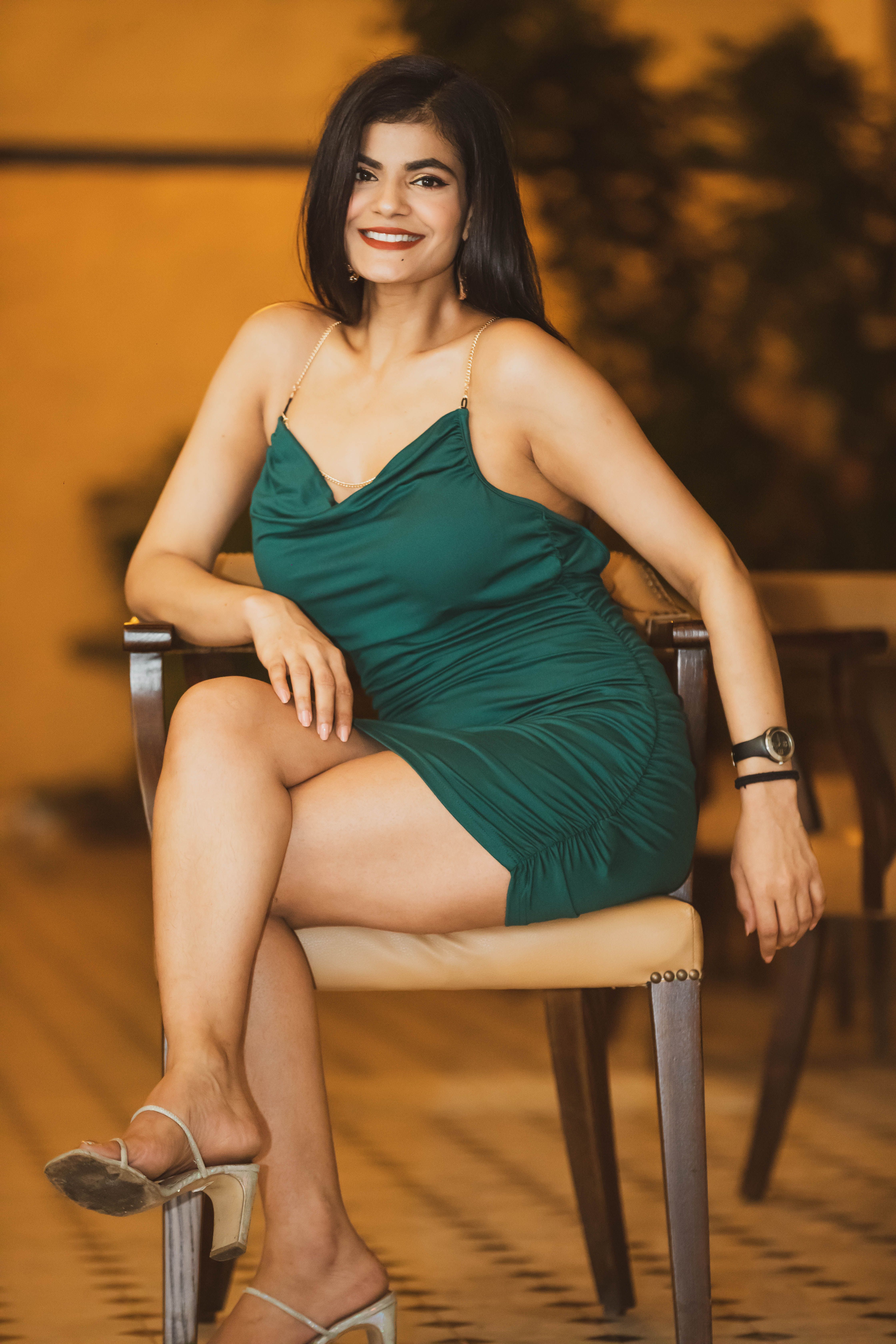 A smiling woman in a green dress seated thoughtfully on a chair indoors.