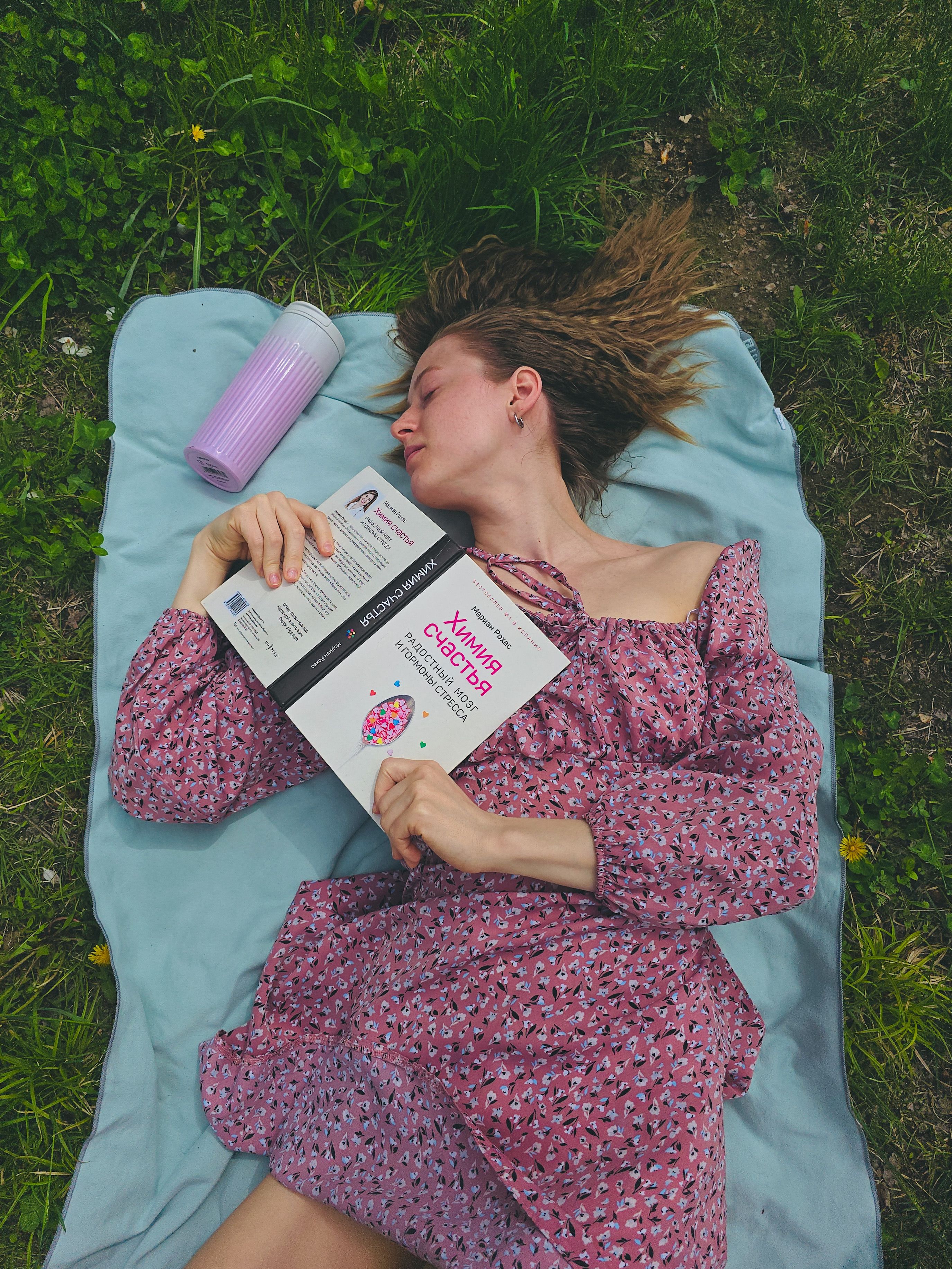 A woman in a floral dress relaxes outdoors on a blanket with a book and drink.