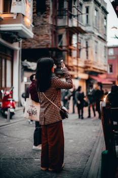A woman photographs an urban street with her smartphone, surrounded by blurred people and buildings.