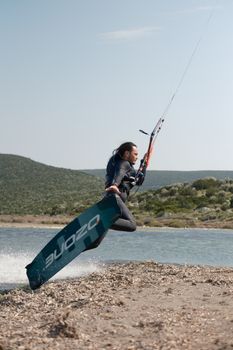 A kiteboarder performs a jump over a tranquil riverside landscape on a sunny day.