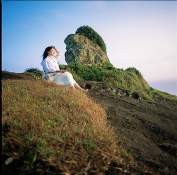 A woman in white sitting on a rocky cliffside, enjoying a peaceful sunset view.