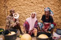 Three African women sitting outdoors preparing traditional food using local tools.