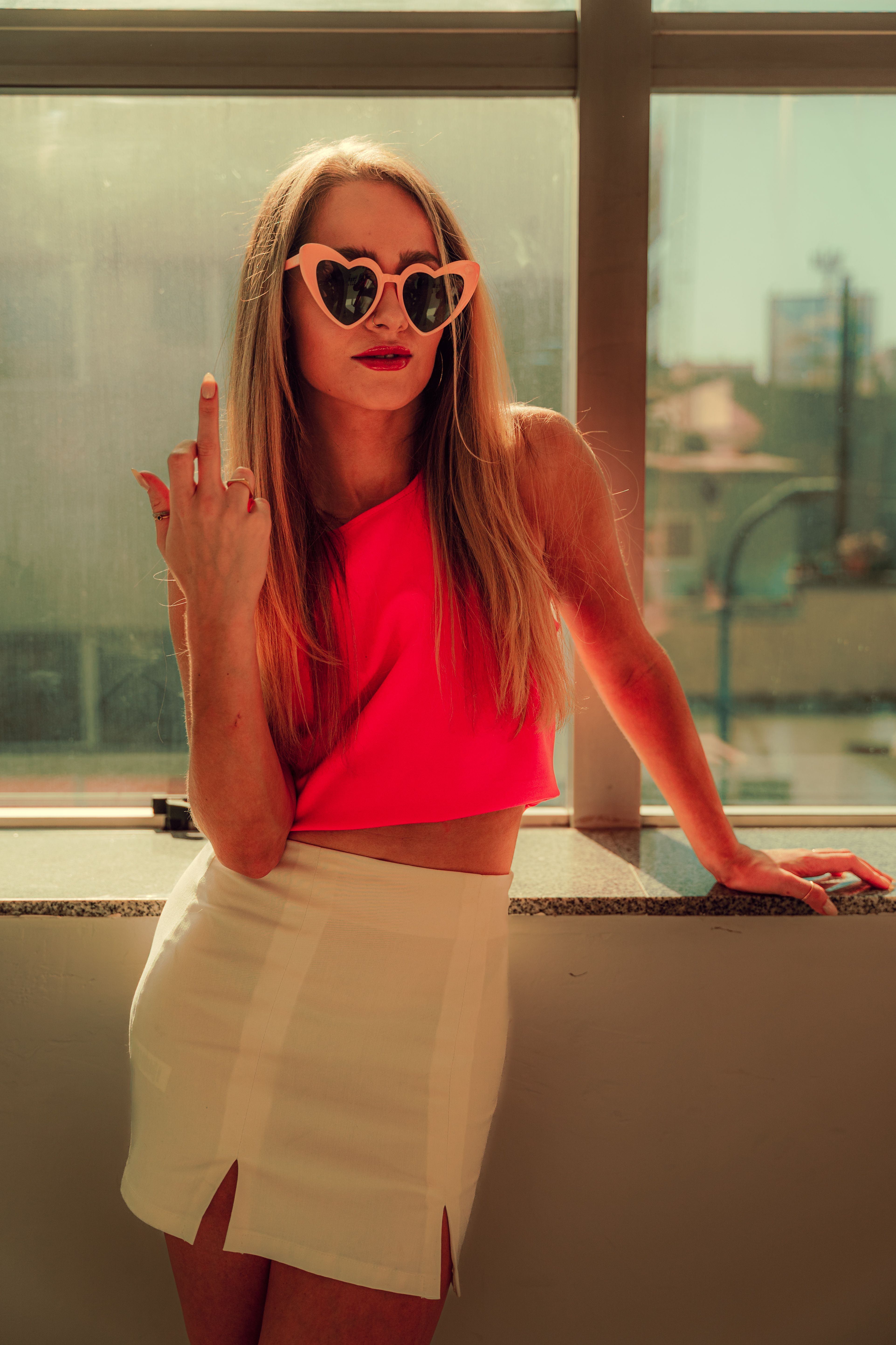 Trendy woman in a pink top and heart sunglasses posing confidently by a window indoors.