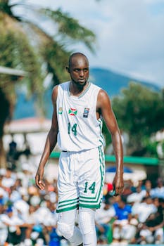 Athlete playing basketball outdoors with palm trees and spectators.