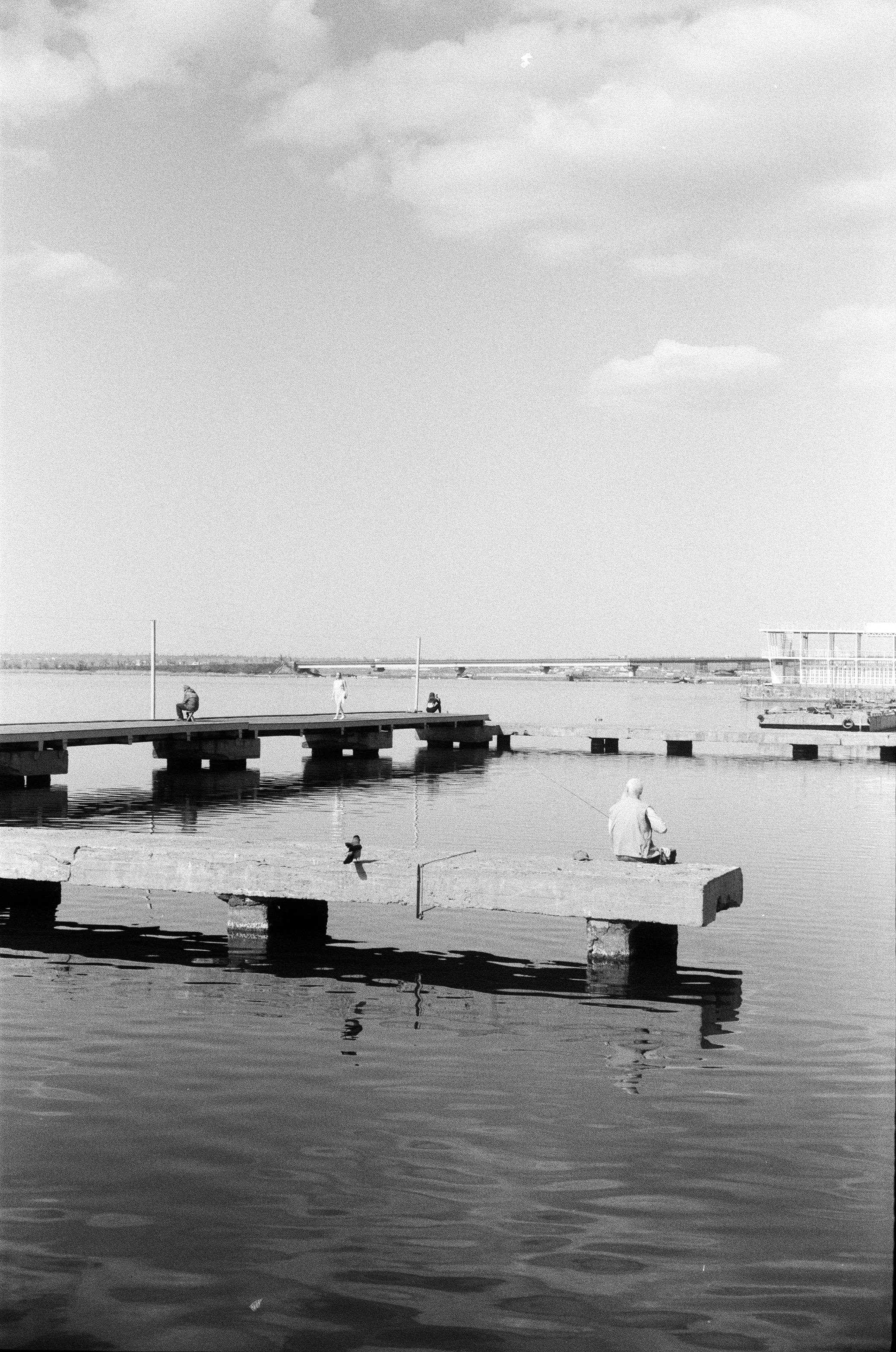 Black and white photo of people relaxing on piers by a lake under a cloudy sky.