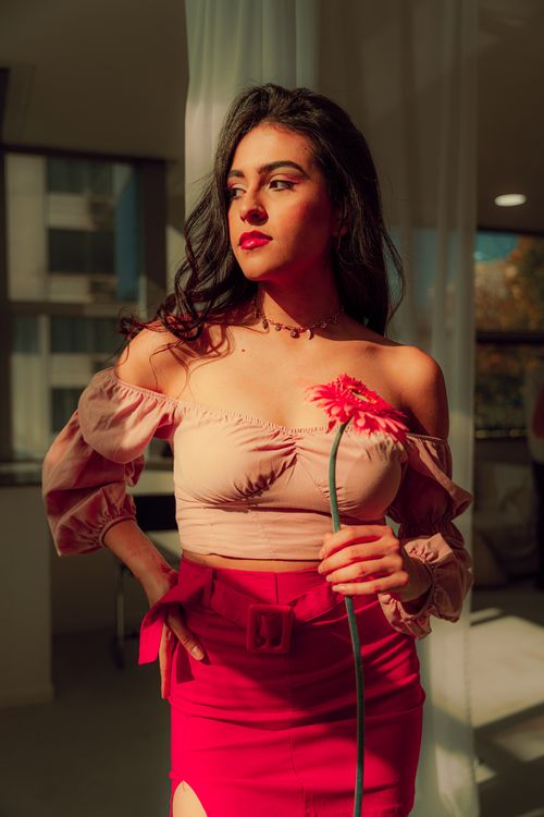 Free Stylish woman in pink outfit holding a red flower in a sunlit room, exuding confidence and grace. Stock Photo