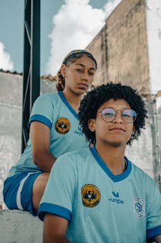 Two young soccer players wearing blue uniforms posing casually outdoors on a sunny day.