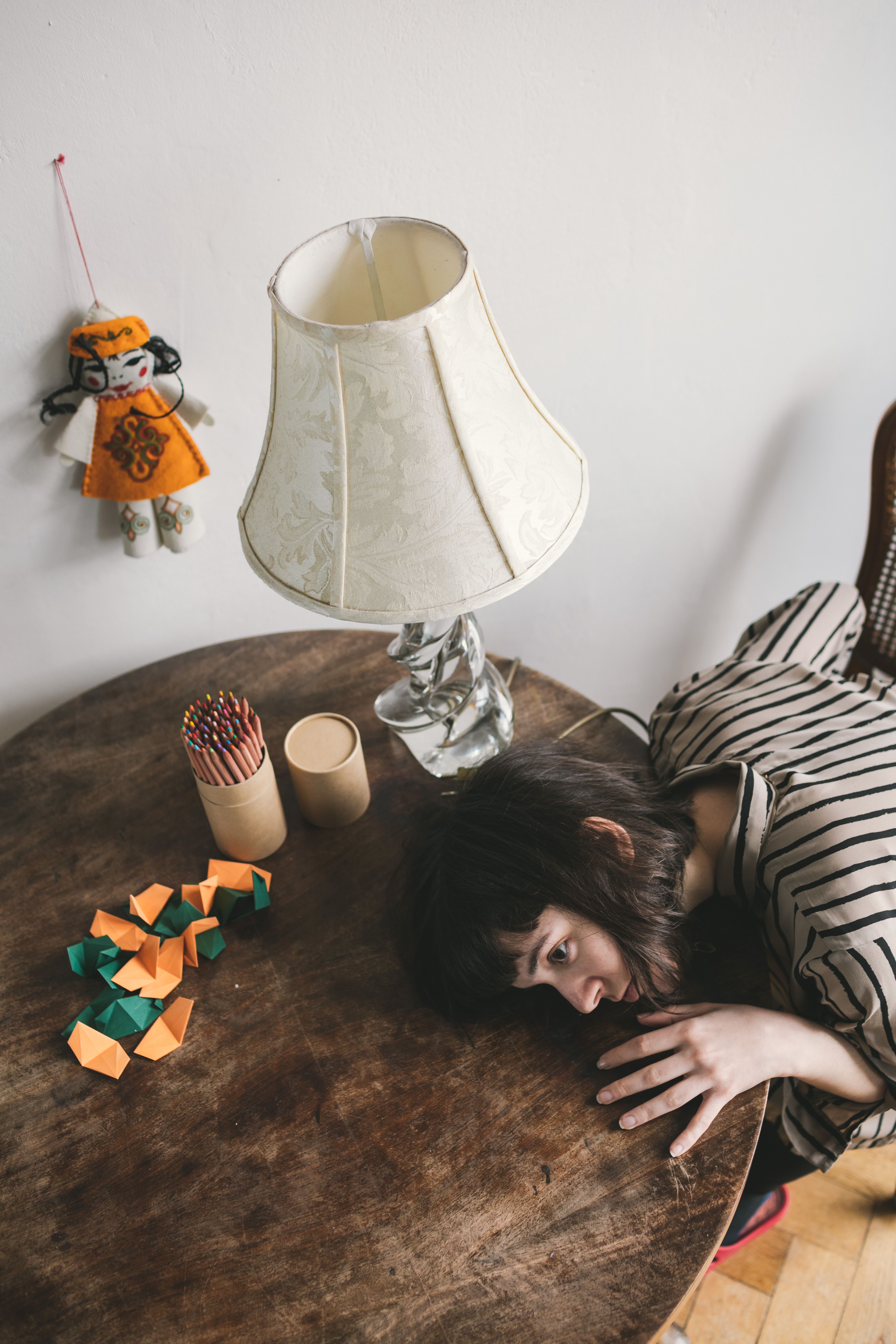 A woman rests her head on a table with colorful origami and a lamp nearby, capturing a serene indoor moment.