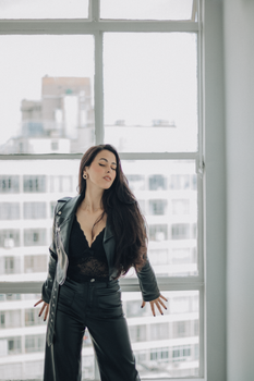 Fashionable woman poses in leather outfit by window with urban backdrop.