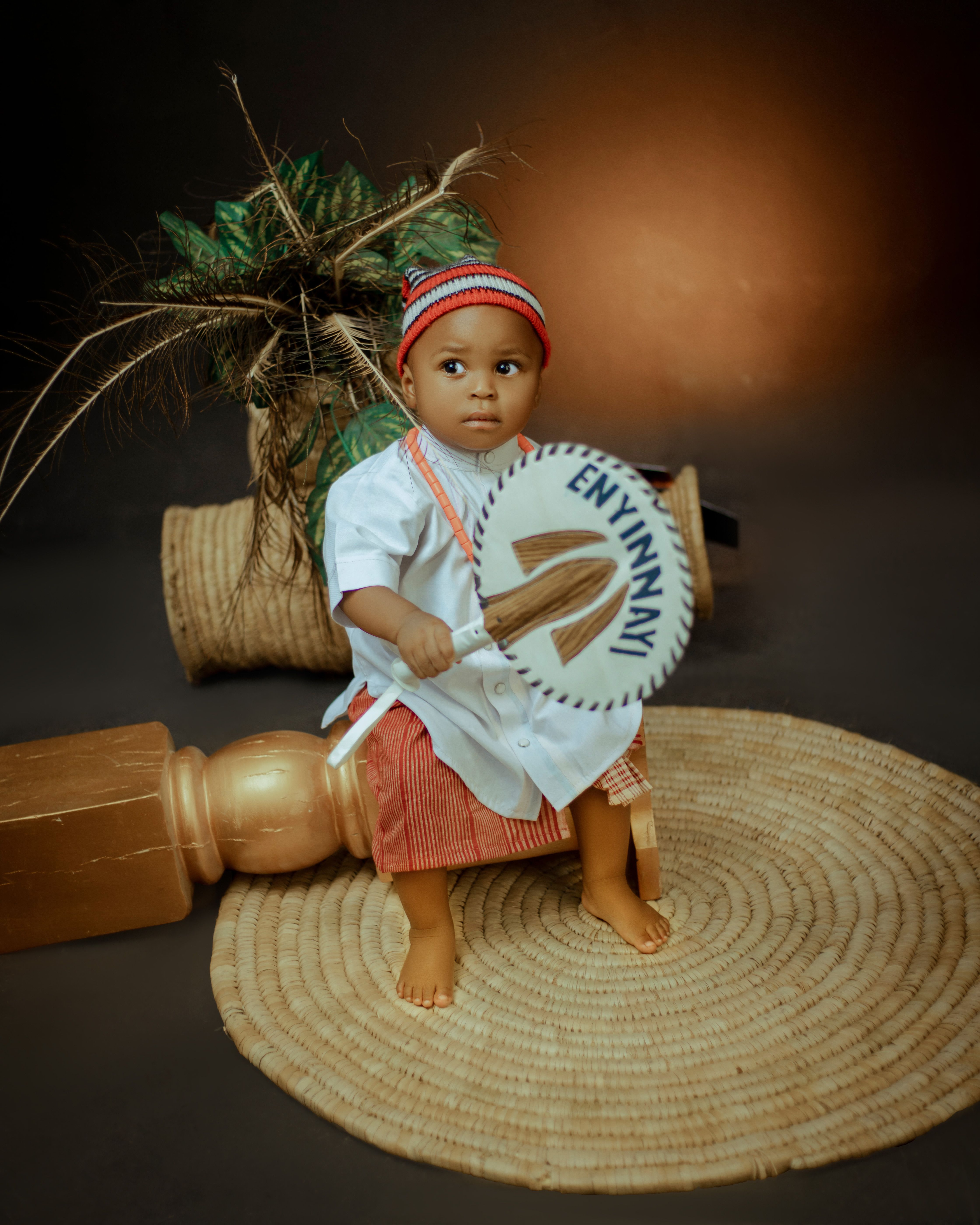 Adorable baby dressed in traditional attire, holding a fan, and surrounded by cultural decor.