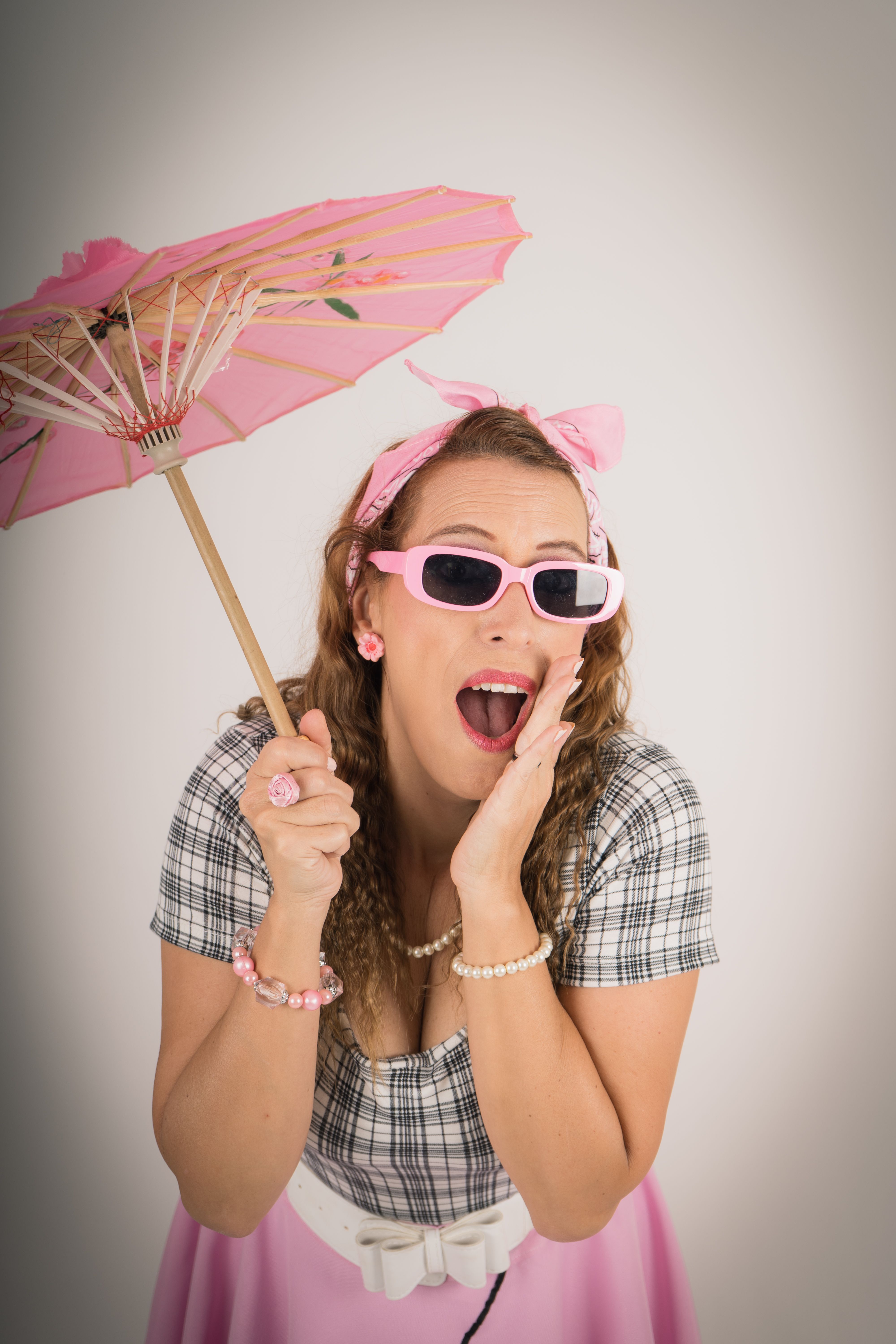 Vintage styled woman with sunglasses and parasol striking a playful pose indoors.