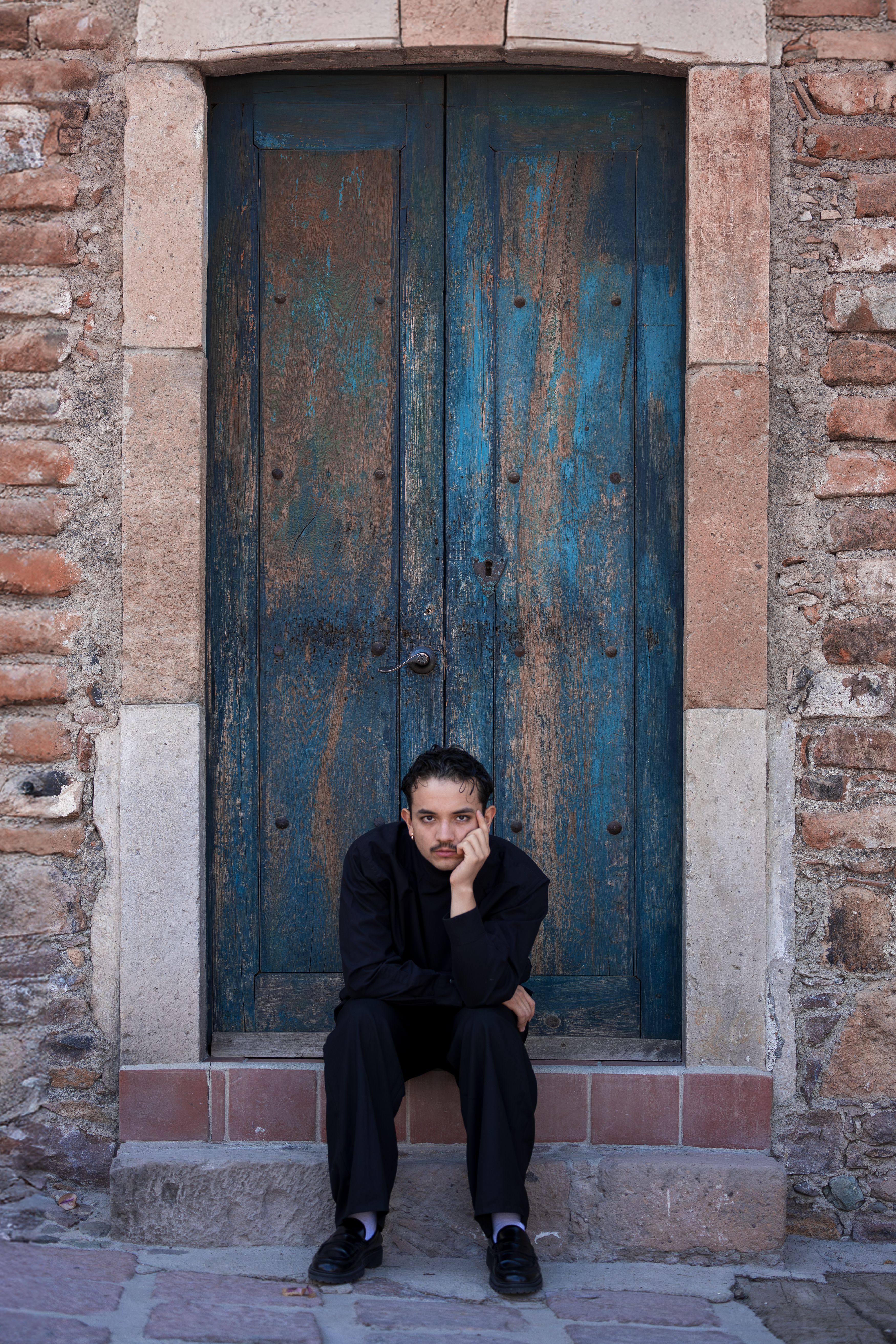 A man in black sits pensively by an old wooden door with rustic bricks.