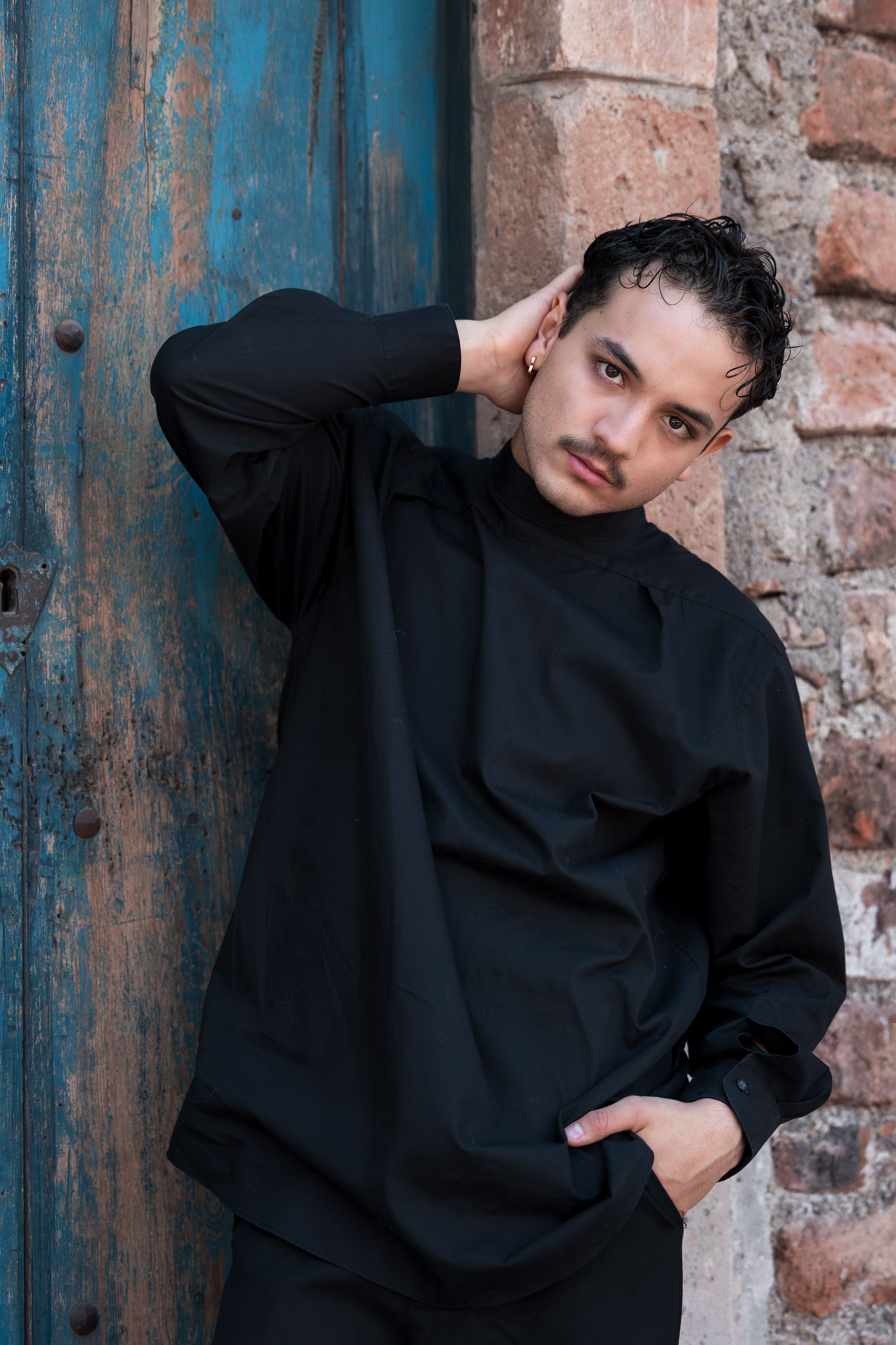 Stylish young man in black outfit posing against a rustic brick wall and wooden door.