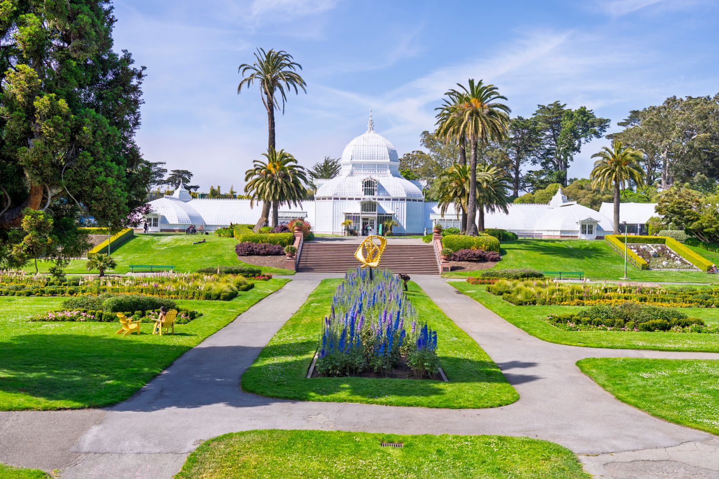 The white Victorian greenhouse of the Conservatory of Flowers in Golden Gate Park, San Francisco, framed by palm trees and colorful flower beds under a sunny sky