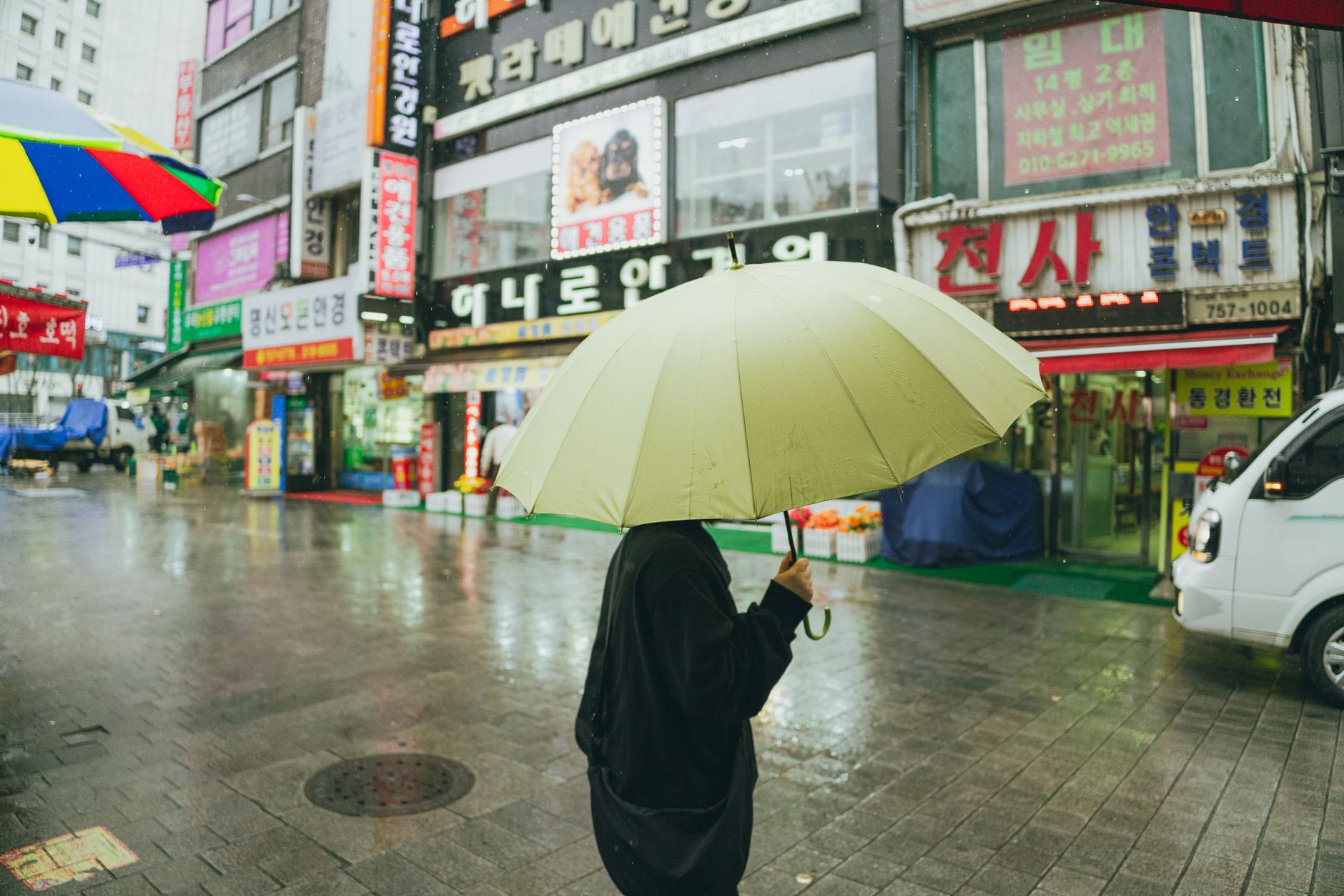 서울 하늘에 가벼운 봄비가 내리는 아침 풍경, 흐린 하늘 아래 녹색 나무와 젖은 거리, 기온 14도 표시된 디지털 간판이 보이는 장면
