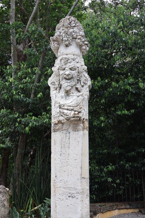 Free Ornate stone column sculpture surrounded by dense greenery in an outdoor garden. Stock Photo