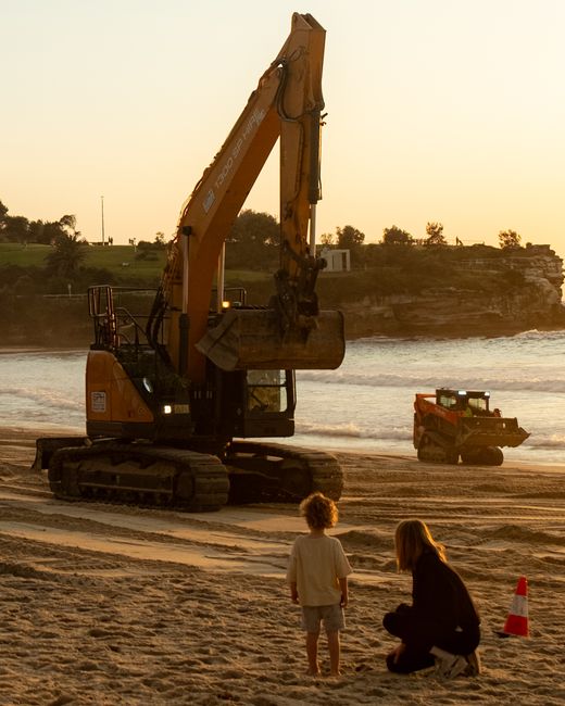 A child and adult by a digger at Coogee Beach during sunset, showcasing construction work.