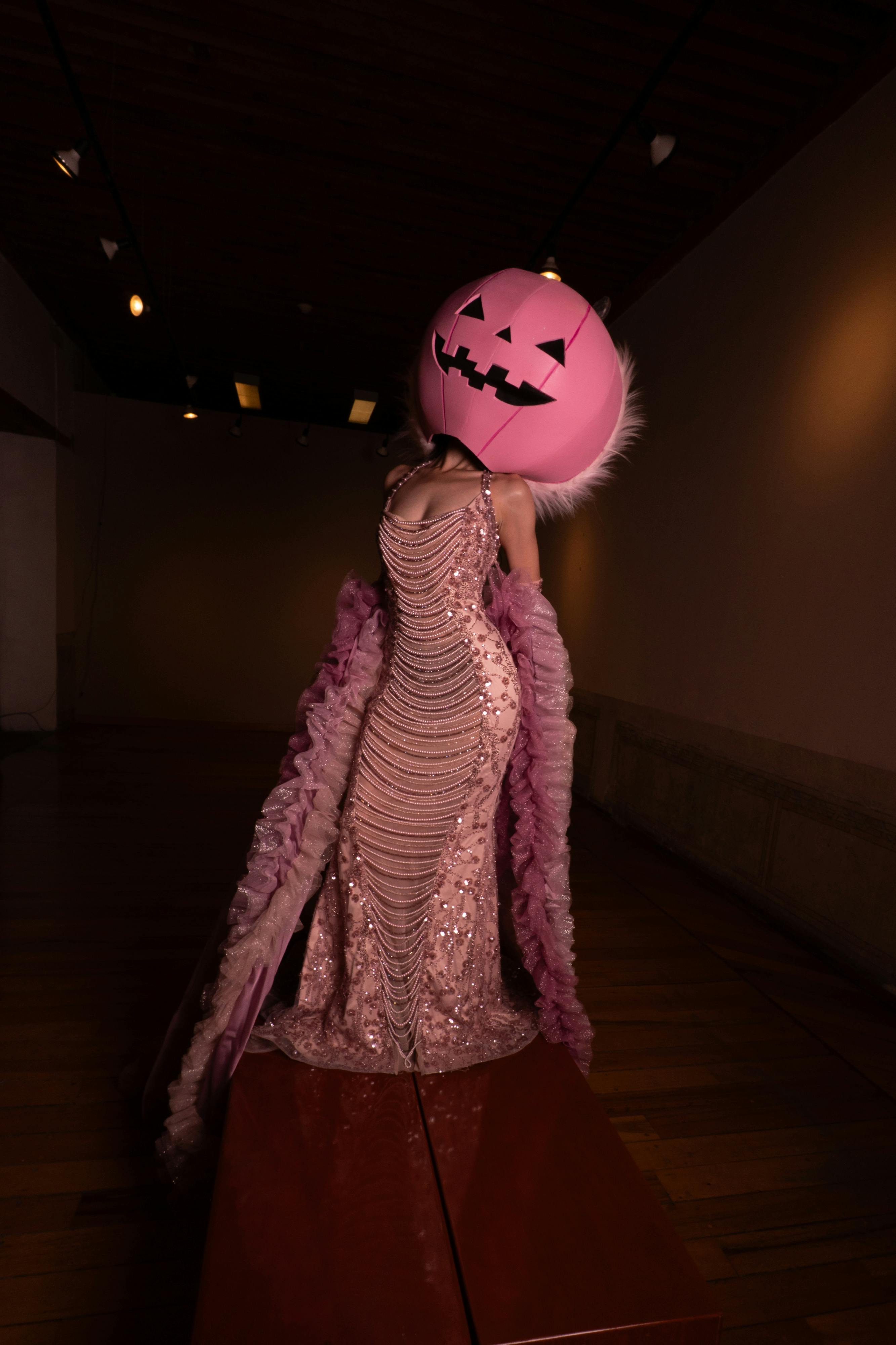 A glamourous pink gown paired with a unique pink pumpkin head in a dimly lit room.