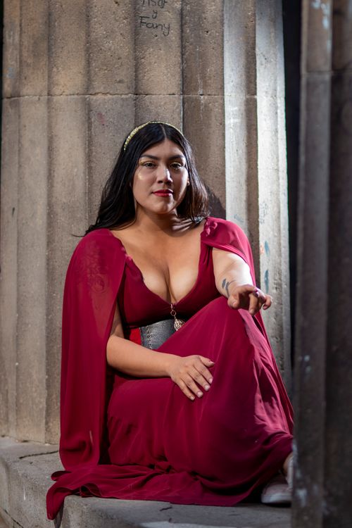 Free An elegant woman in a flowing red gown sits gracefully against ancient stone pillars. Stock Photo