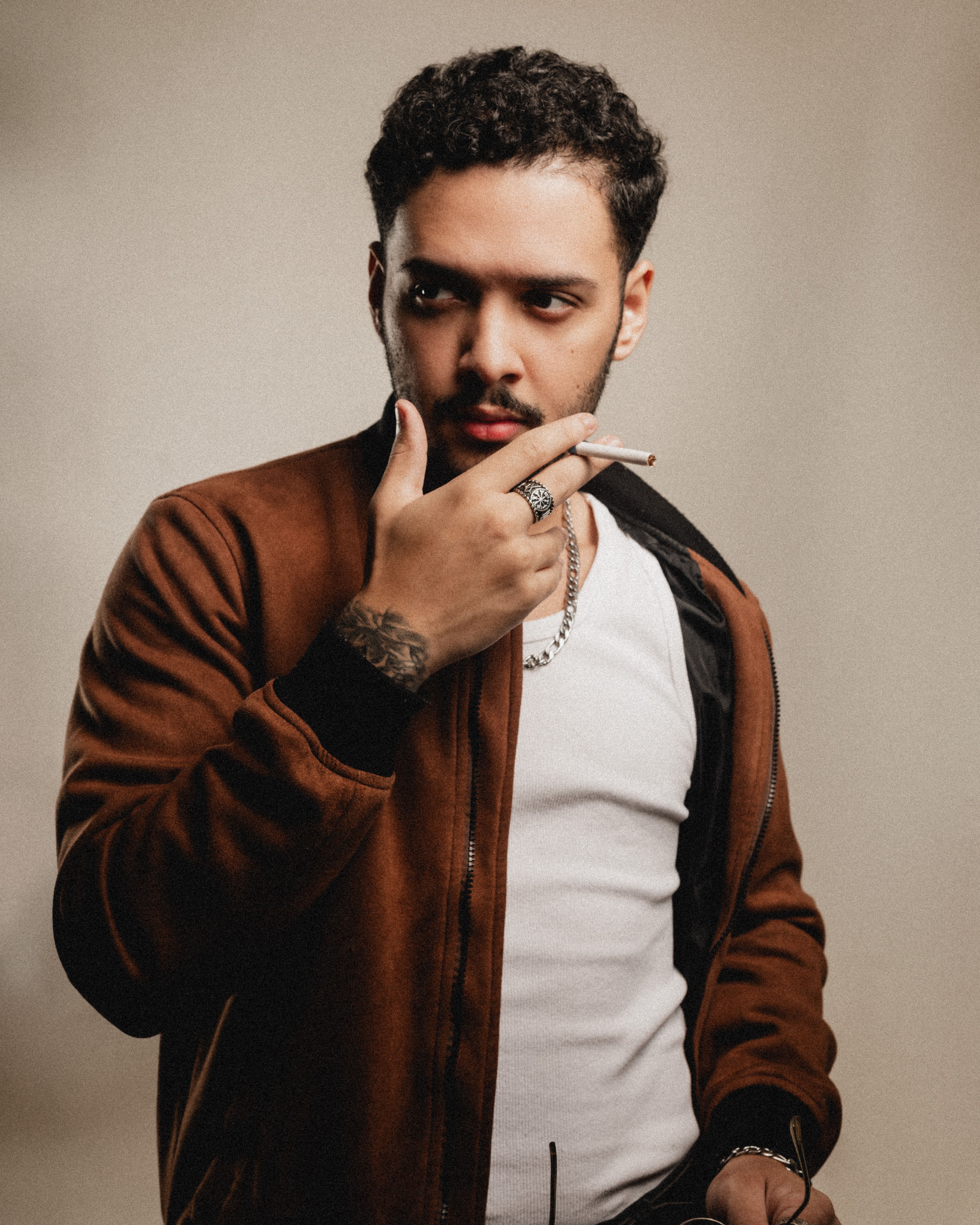A man in a casual jacket poses thoughtfully while holding a cigarette, expressing a moody and stylish vibe.