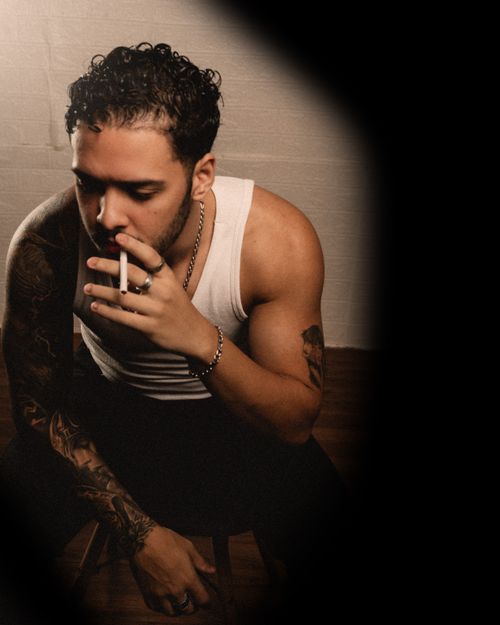 Free A young man with tattoos sits indoors in a contemplative pose, showcasing a moody atmosphere. Stock Photo